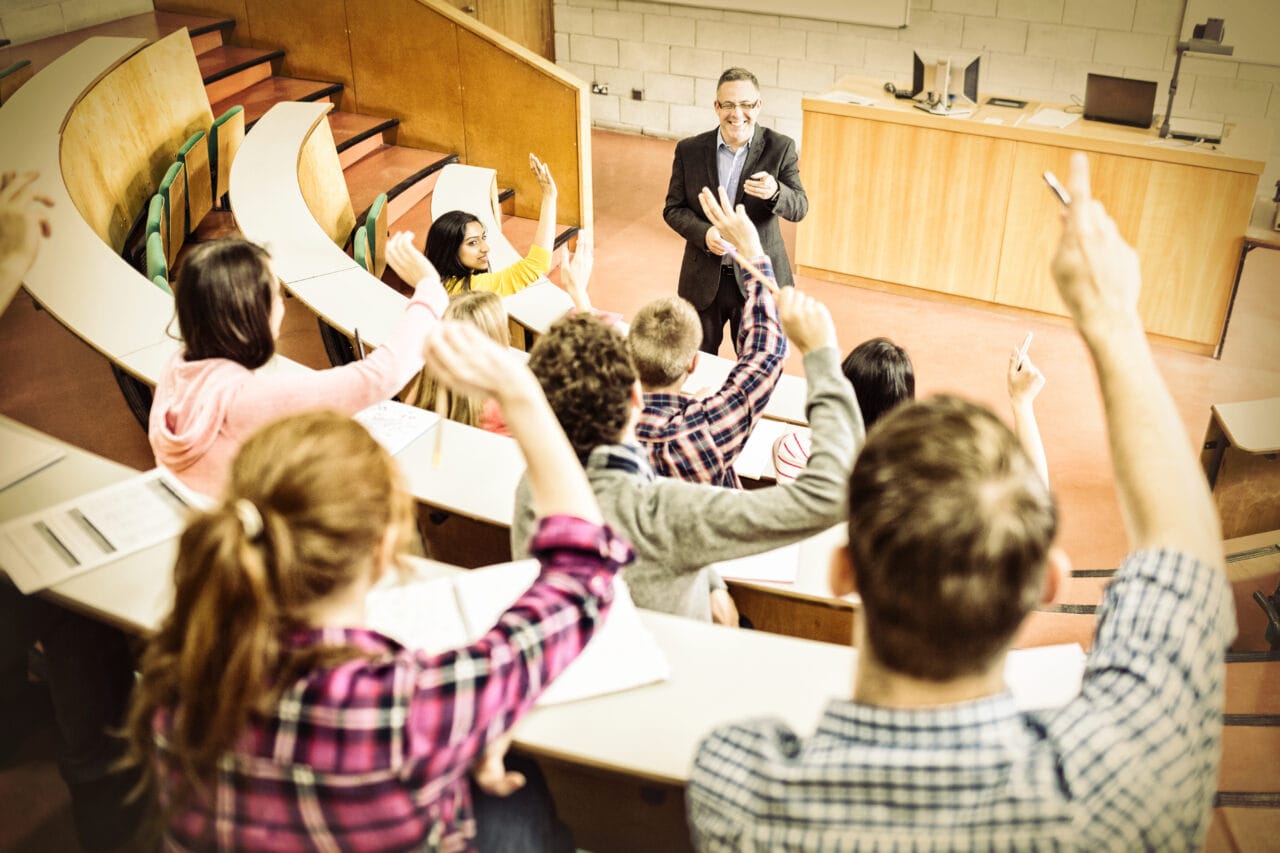 Studenci w sali wykładowej podnoszący ręce, profesor prowadzący zajęcia przy mównicy.